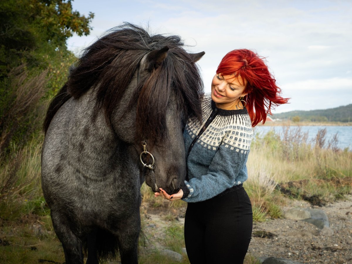 Rödhårig kvinna i vacker stickad tröja och en svart islandshäst vid havet (fotograf Hillevi Upmanis)
